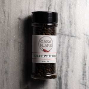 A 3 oz clear plastic spice jar of Casa Flake Black Peppercorns with a black lid and white label, set against a gray marble background.