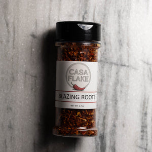 A clear plastic spice jar of Casa Flake Blazing Roots seasoning with a black lid and white label, set against a gray marble background.