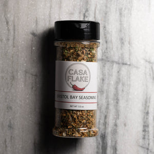 A clear plastic spice jar of Casa Flake Bristol Bay Seasoning with a black lid and white label, set against a gray marble background.