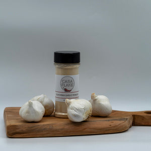 Bottle of Casa Flake garlic powder on a wooden cutting board with garlic bulbs against a gray background