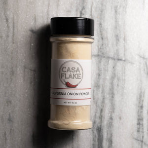 A clear plastic spice jar of Casa Flake California Onion Powder with a black lid and white label, set against a gray marble background.