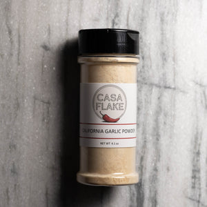 A bottle of Casa Flake California Garlic Powder with a black lid and white label, set against a gray marble background.
