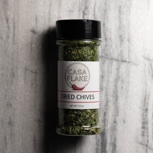 A clear plastic spice jar of Casa Flake Dried Chives with a black lid and white label, set against a gray marble background.