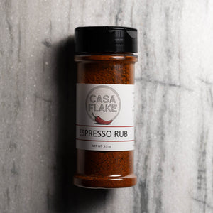 A clear plastic spice jar of Casa Flake Espresso Rub with a black lid and white label, set against a gray marble background.
