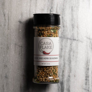 A clear plastic shaker bottle of Casa Flake Garlic and Herb Seasoning with a black lid and white label, set on a gray marble surface.