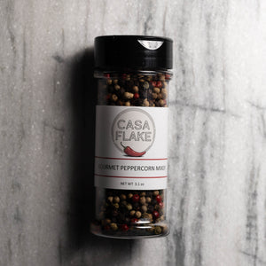 A clear plastic spice jar of Casa Flake Gourmet Peppercorn Mix with a black lid and white label, set against a gray marble background.