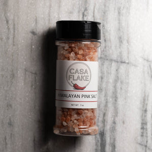 A 7 oz clear plastic shaker bottle of Casa Flake Himalayan Pink Salt with a black lid and white label, set against a gray marble background.