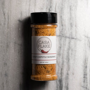 A clear plastic spice jar filled with orange-brown seasoning, topped with a black lid and labeled CASA FLAKE Honey Chipotle Seasoning, set against a gray marble-textured background.