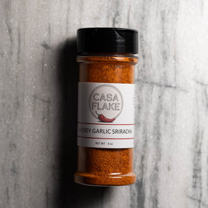 A bottle of Casa Flake Honey Garlic Sriracha seasoning, showing the orange-brown spice powder inside, a white label with the brand logo, and a black cap, set against a gray marble background.