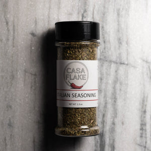 A clear plastic spice jar filled with Italian seasoning, featuring a white label with the brand name 'CASA FLAKE' and a black lid, set against a gray marble background.