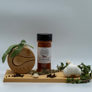 Casa Flake KC BBQ Rub jar on a wooden board with herbs and spices on a light gray background