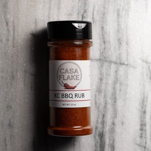 A bottle of Casa Flake KC BBQ Rub seasoning standing upright on a gray stone-textured surface. The clear bottle shows the reddish-brown spice inside, with a white label and black cap.