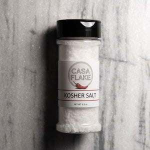 A clear plastic shaker bottle of Casa Flake Kosher Salt with a black flip-top lid and a white label, set against a gray marble-textured background.