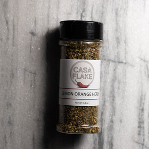 A clear plastic spice jar with a black lid and a white label reading 'CASA FLAKE' and 'LEMON ORANGE HERBS', filled with dried greenish-brown herbs, photographed on a gray marble-like surface.