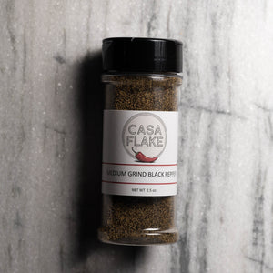 A clear plastic shaker bottle of Casa Flake Medium Grind Black Pepper seasoning, featuring a white label with a red chili pepper graphic and a black lid, set against a gray marble background.