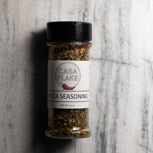 A clear plastic spice jar of Casa Flake Pizza Seasoning with a black lid and white label, set against a gray marble background.