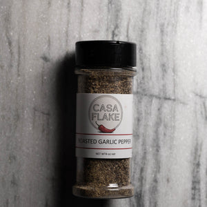 A clear plastic spice jar of Casa Flake Roasted Garlic Pepper with a black lid and white label, set against a gray marble-textured background.