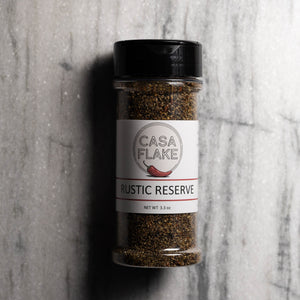 A clear plastic spice jar of Casa Flake Rustic Reserve seasoning with a black lid and white label, set against a gray marble background.