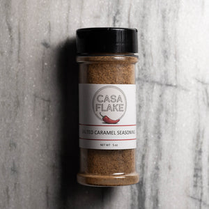 A 5 oz clear plastic spice jar of Casa Flake Salted Caramel Seasoning with a black lid and white label, set against a gray marble background.
