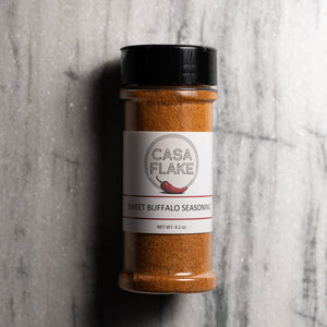 A clear plastic spice jar filled with orange-brown seasoning powder, topped with a black lid and labeled 'CASA FLAKE SWEET BUFFALO SEASONING,' set against a gray stone-textured background.