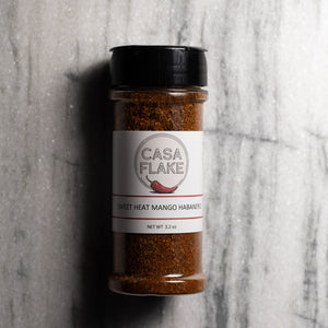 A 3.2 oz shaker bottle of Casa Flake Sweet Heat Mango Habanero seasoning, featuring a white label and black cap, set against a gray marble background.